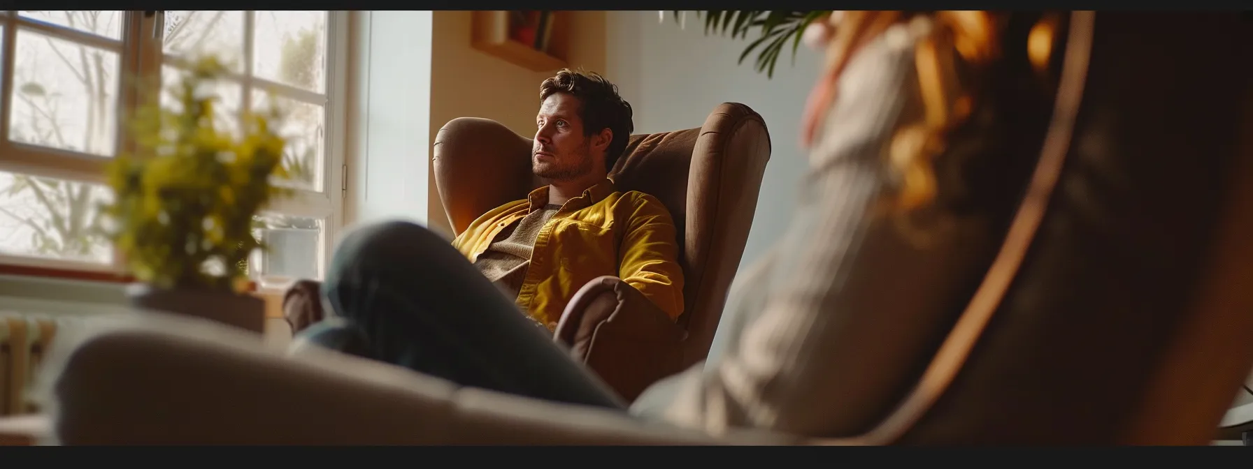 a person sitting in a comfortable chair during a therapy session, while a therapist listens attentively.